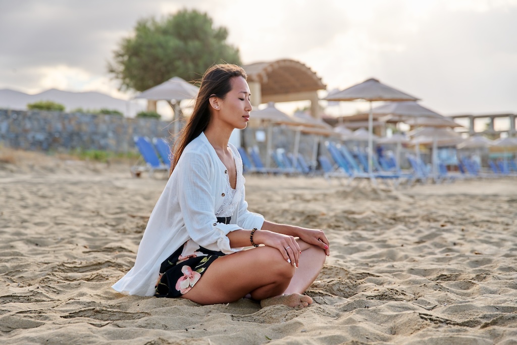 Young relaxed woman sitting in lotus position on beach, morning sea nature background. Natural Asian woman with closed eyes on sand in Southern California.